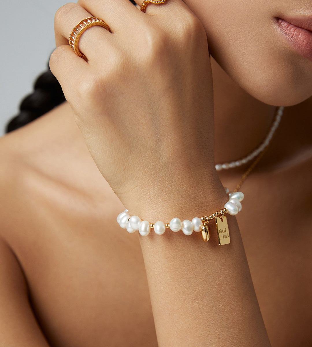 Good Luck baroque pearl bracelet worn on wrist in vintage gold — "Good Luck" tag and heart charm detail, styled with gold rings and pearl necklace, 18K gold plated S925 silver, BUCKLE Collective