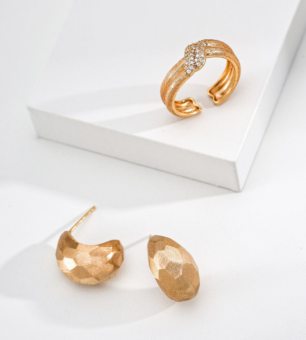 A pair of 18K gold-plated S925 silver textured crescent-shaped hoop earrings and a matching gold-tone wrap ring with a row of clear stones, displayed on a clean white surface with a white geometric block.