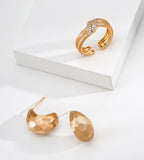 A pair of 18K gold-plated S925 silver textured crescent-shaped hoop earrings and a matching gold-tone wrap ring with a row of clear stones, displayed on a clean white surface with a white geometric block.