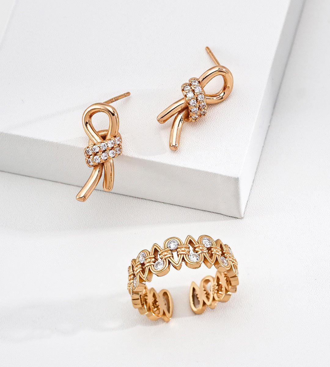 An 18K gold-plated S925 silver jewelry set featuring a bow-shaped stud earring with a crystal-encrusted center and a matching open ring with interlocking bow motifs and clear stones, displayed on a white geometric surface.