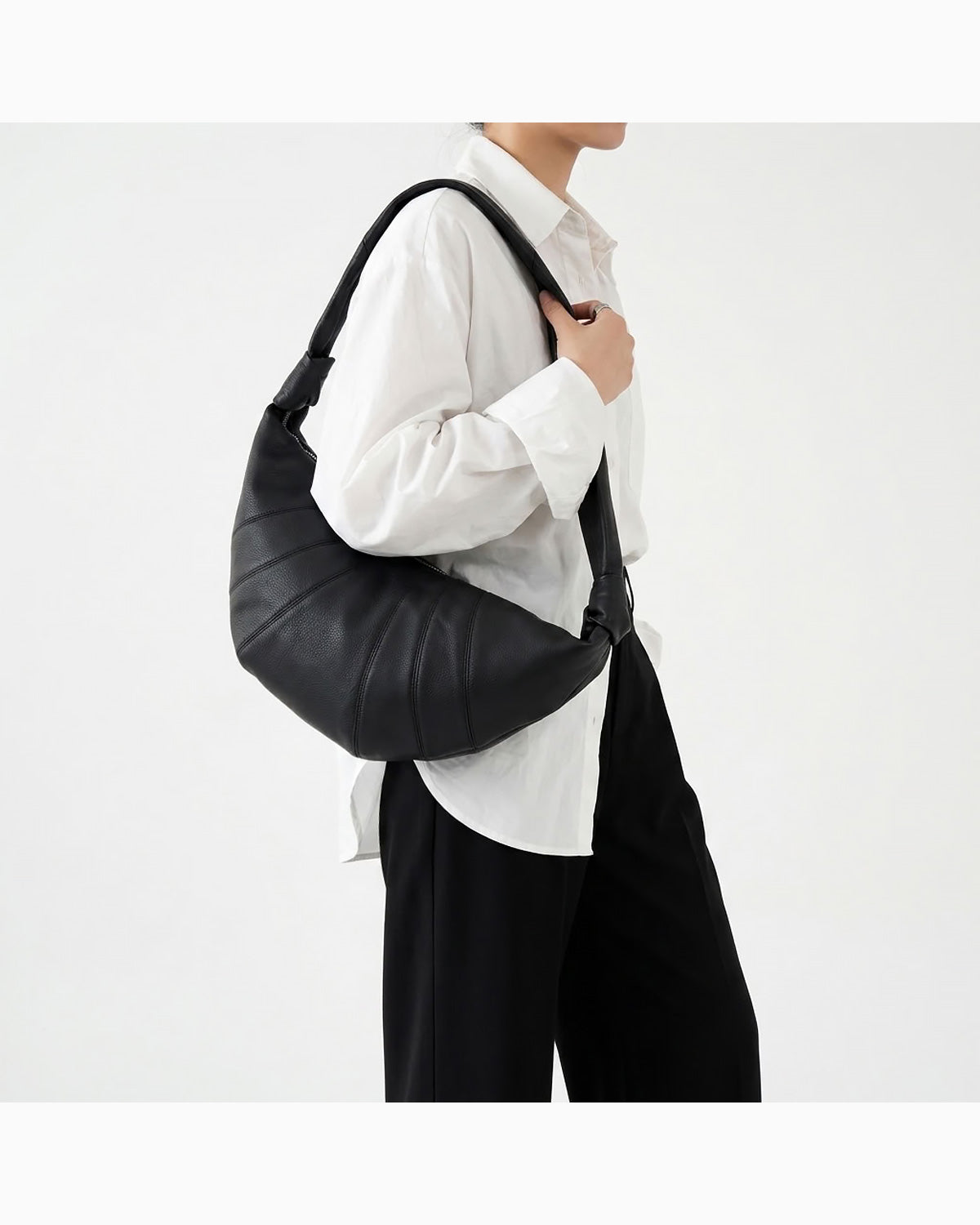 A woman wearing a white button-up shirt and black pants carries a large black crescent-shaped lambskin shoulder bag by BUCKLE Collective