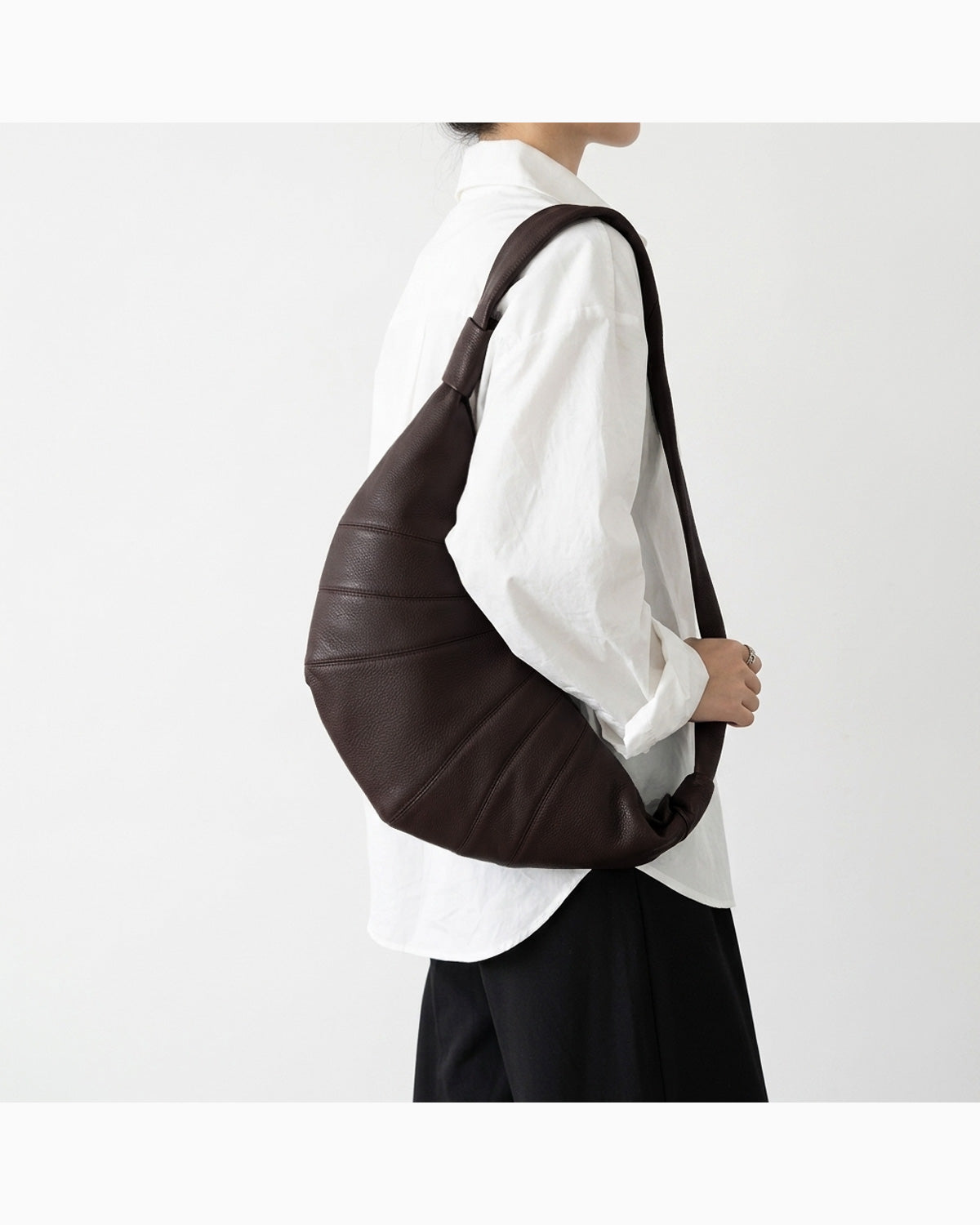 A person wearing a white shirt and black pants carries a large Coffee-brown Top-grain Lambskin Leather Croissant Crossbody Bag | BUCKLE Collective