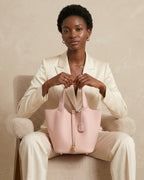 A woman in a cream-colored blazer and trousers sits on a plush beige armchair, holding a light pink leather bucket bag with gold-tone hardware.