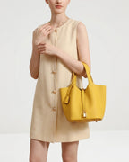A model wearing a beige sleeveless button-front dress and carrying a Large Yellow Genuine Togo Leather Bucket Bag with a gold lock