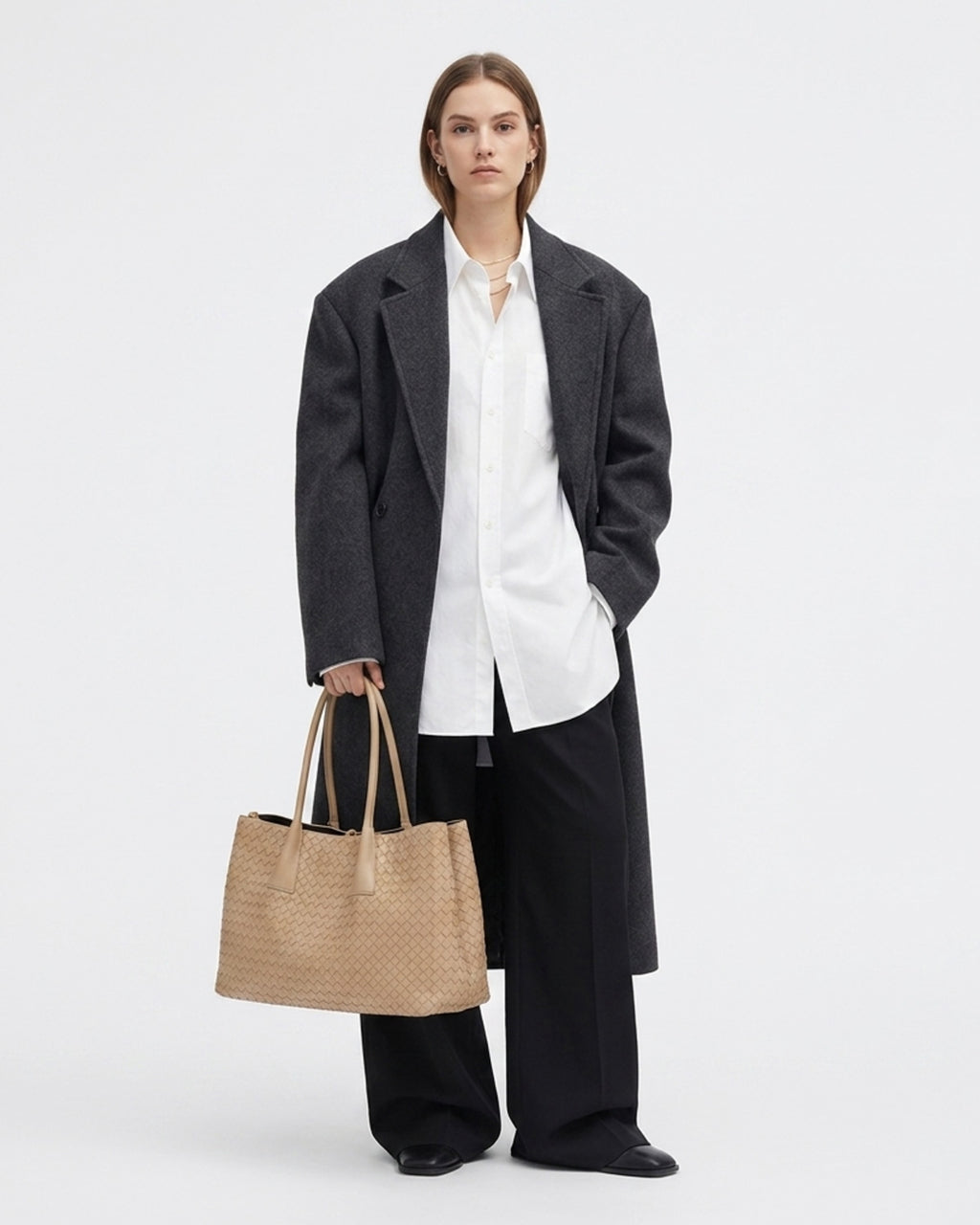 A model wearing an oversized dark charcoal coat over a white button-down shirt and black wide-leg trousers, while holding a beige woven lether tote bag.