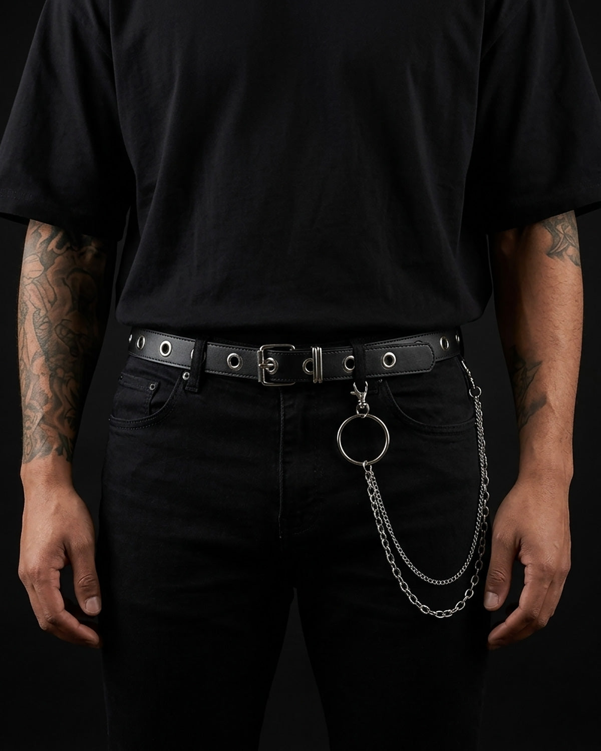 A black leather belt with silver hardware, worn by a man. The belt features a silver buckle, silver eyelets, and a prominent silver O-ring with two hanging chains, styled with black jeans and a black t-shirt | BUCKLE Collective