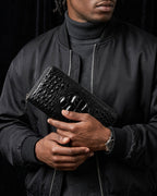 A man wearing a black bomber jacket and gray turtleneck holds a Genuine Siamese Crocodile Leather Clutch