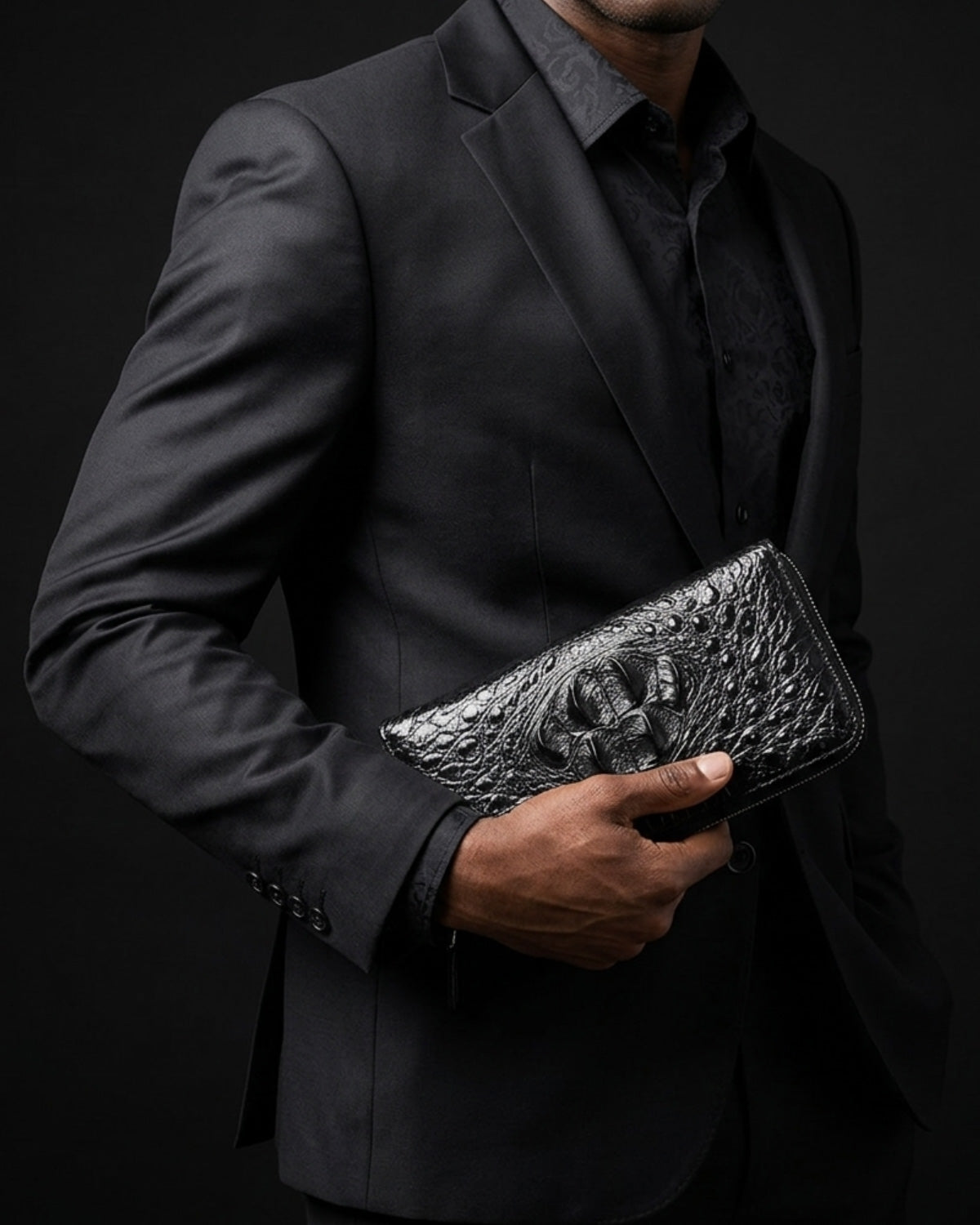 A man in a black suit and shirt holding a black crocodile leather clutch