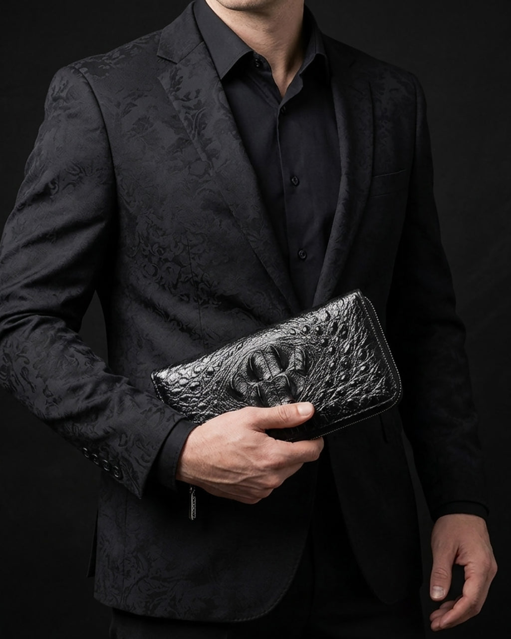 A man in a black patterned suit jacket and black shirt holds a black crocodile leather clutch