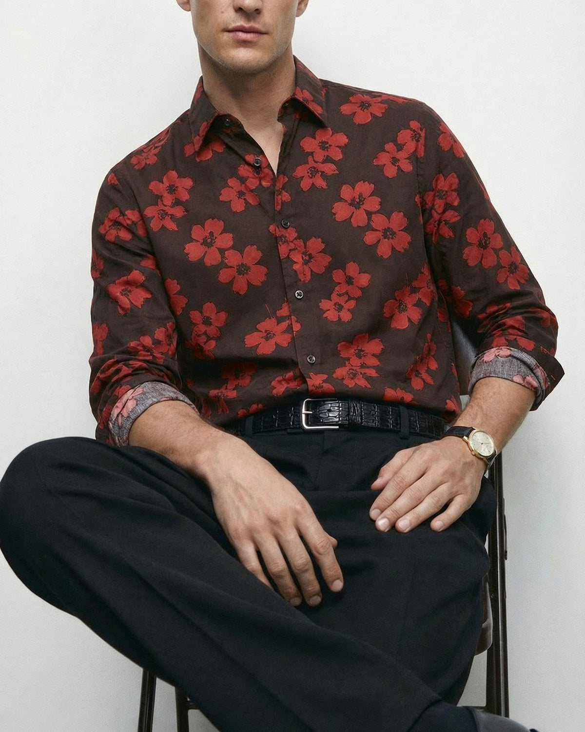 A man wearing a dark brown long-sleeve button-up shirt with a red floral pattern, paired with black trousers and a black leather crocodile-pattern belt, seated against a light gray background.