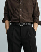 A person wearing a dark brown long-sleeve button-up shirt, black pleated trousers, and a men's black textured leather belt with a silver buckle, shown from the chest down against a light gray background.