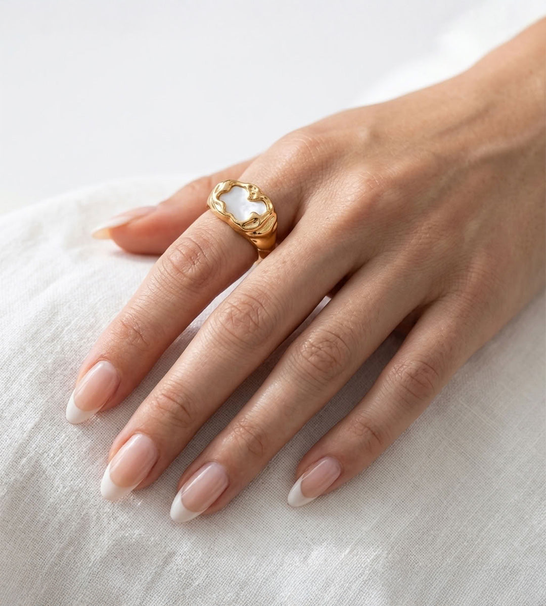Molten mother-of-pearl ring with an irregular gold-tone frame, worn on the index finger against white linen.