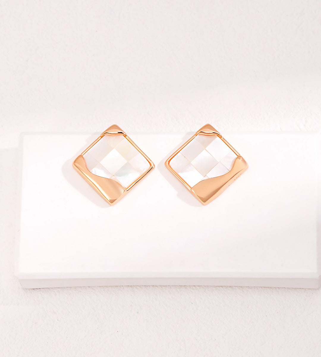 A pair of geometric diamond-shaped stud earrings with an 18K gold-plated finish and a mother-of-pearl center, displayed on a white rectangular block against a textured white background.