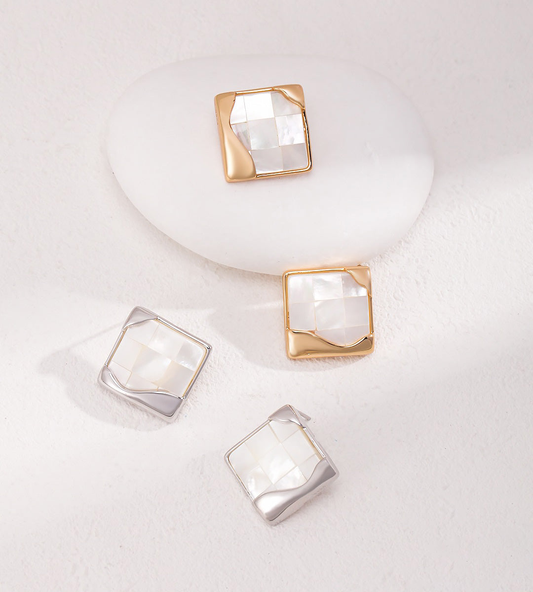 Two pairs of square stud earrings with faceted white mother-of-pearl centers and 18K gold-plated bezel settings, arranged on a white textured surface with one earring displayed on a white oval stand.
