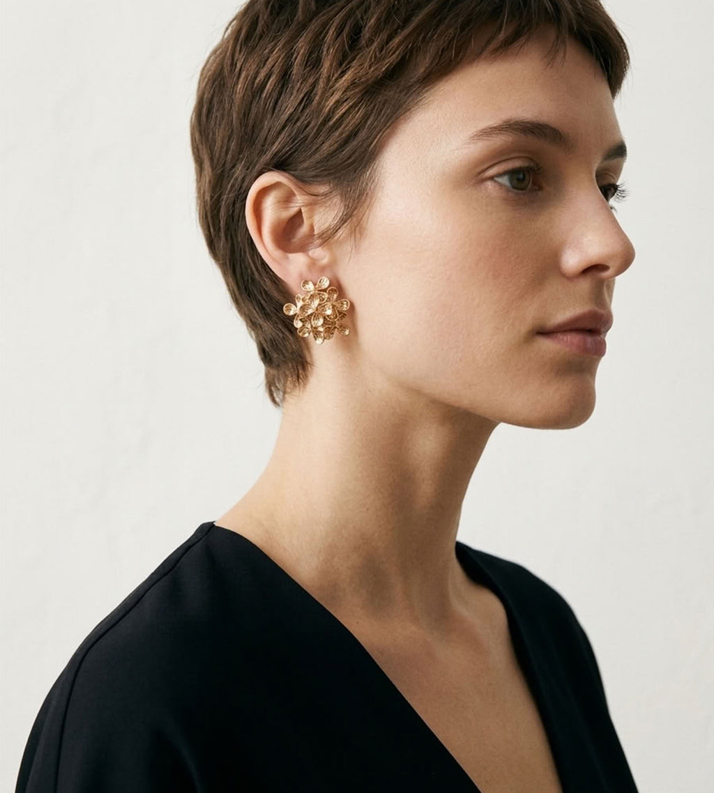 A woman in profile wearing a pair of chunky gold-toned floral cluster earrings, shown against a plain light background.