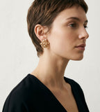 A woman in profile wearing a pair of chunky gold-toned floral cluster earrings, shown against a plain light background.