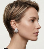 A close-up side profile of a woman with a short brown hairstyle wearing a sparkling silver-toned earring with clear stones against a light gray background.