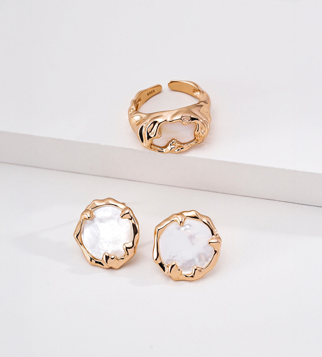 Gold-tone mother-of-pearl open ring displayed above a pair of round shell stud earrings on white blocks.