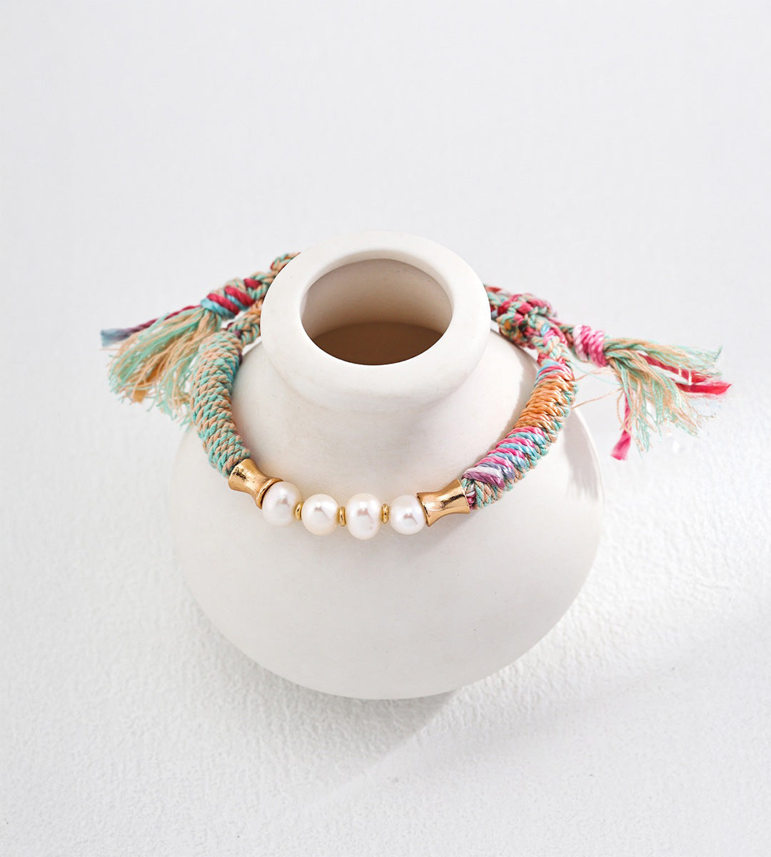 Natural pearl ethnic style adjustable braided bracelet on white ceramic display — freshwater pearls with bamboo-style 18K gold connectors, multicolour hemp cord, frayed ends, BUCKLE Collective