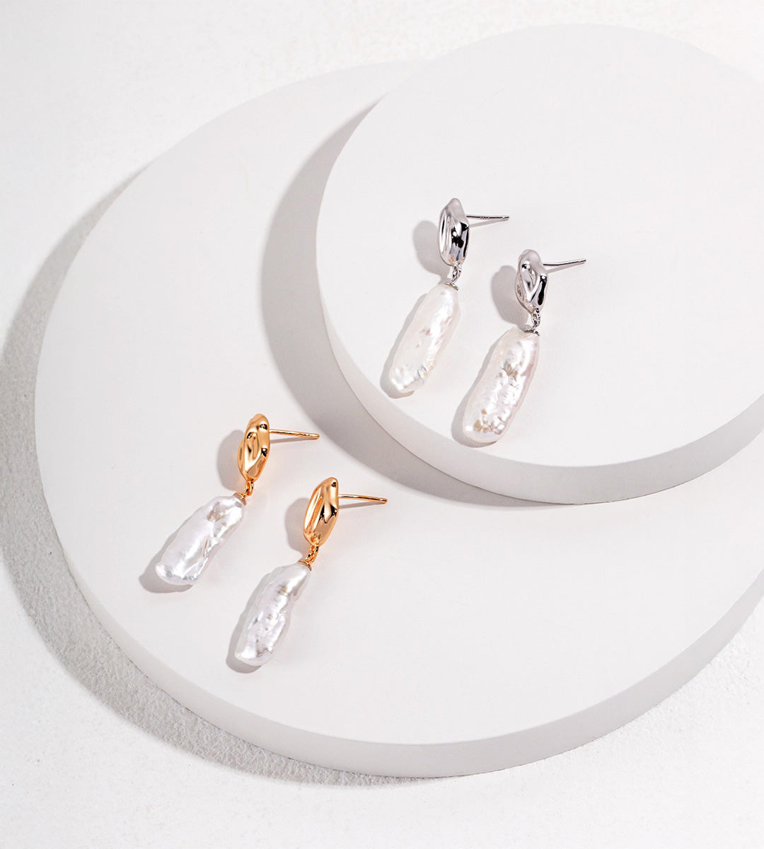 Baroque freshwater pearl drop earrings in Vintage Gold and White Gold finishes on stacked circular white displays.