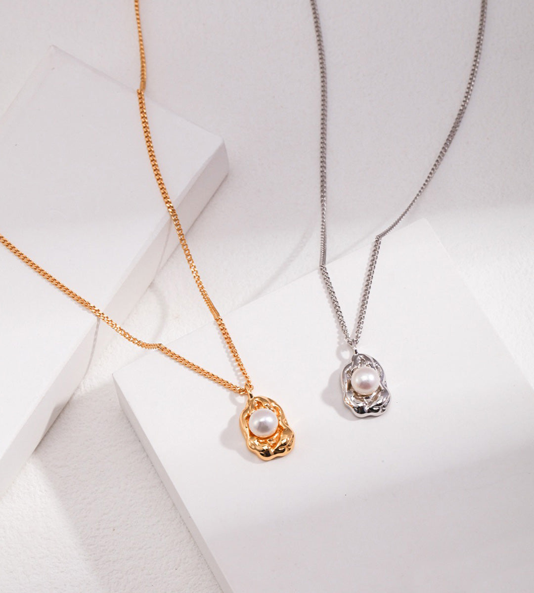 Two delicate pendant necklaces displayed on white geometric blocks: one in warm gold with a white pearl, and one in silver with a white pearl, on a clean white background.