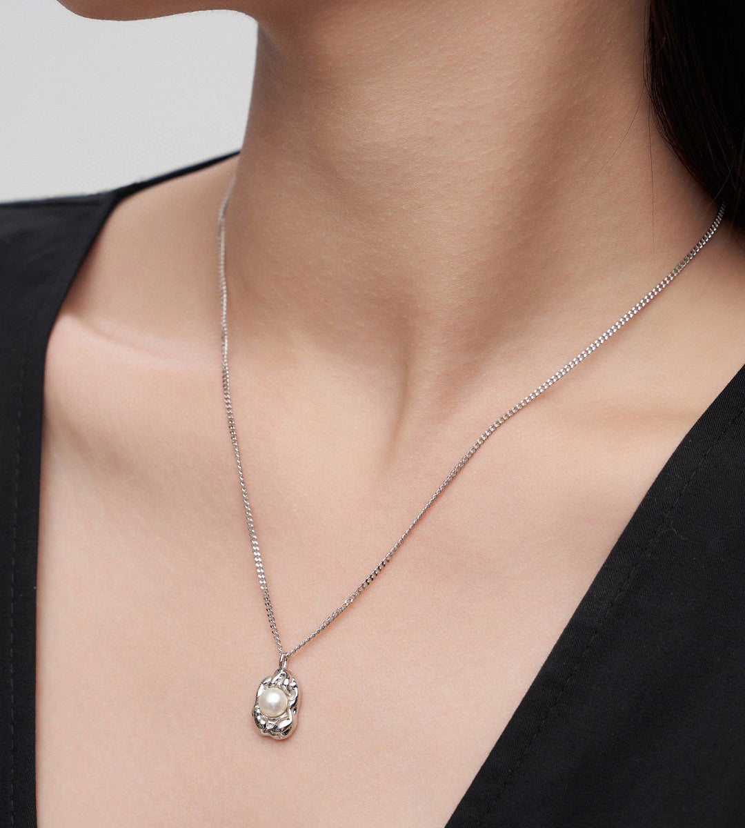 A close-up of a model's neck and chest wearing a delicate 18K white-gold-plated s925 silver chain necklace with an organic-shaped oval pendant featuring a white freshwater pearl in the center