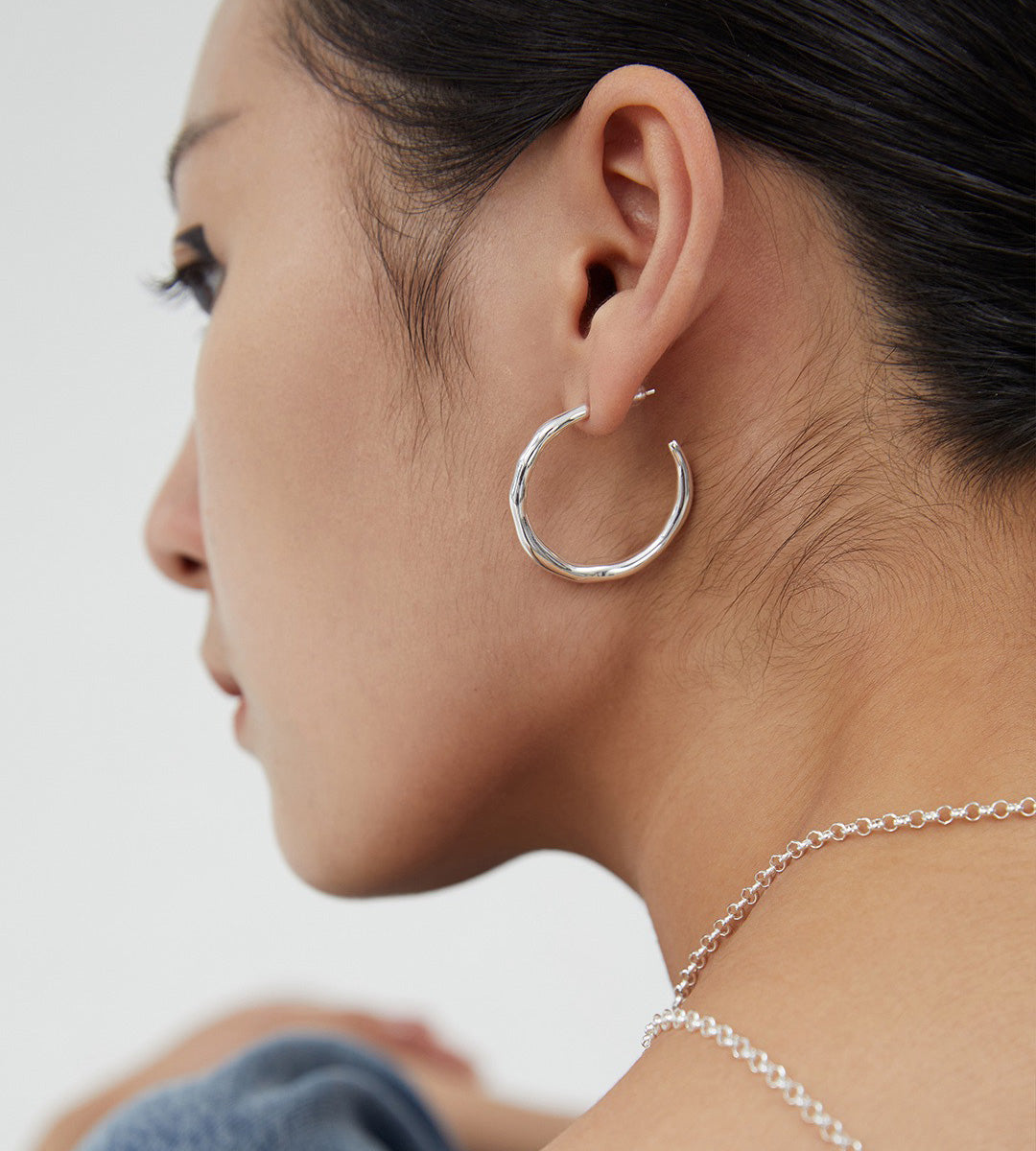 A close-up side profile of a woman wearing a textured 18K white-gold-plated S925 silver hoop earring and a matching silver chain necklace against a plain light background.