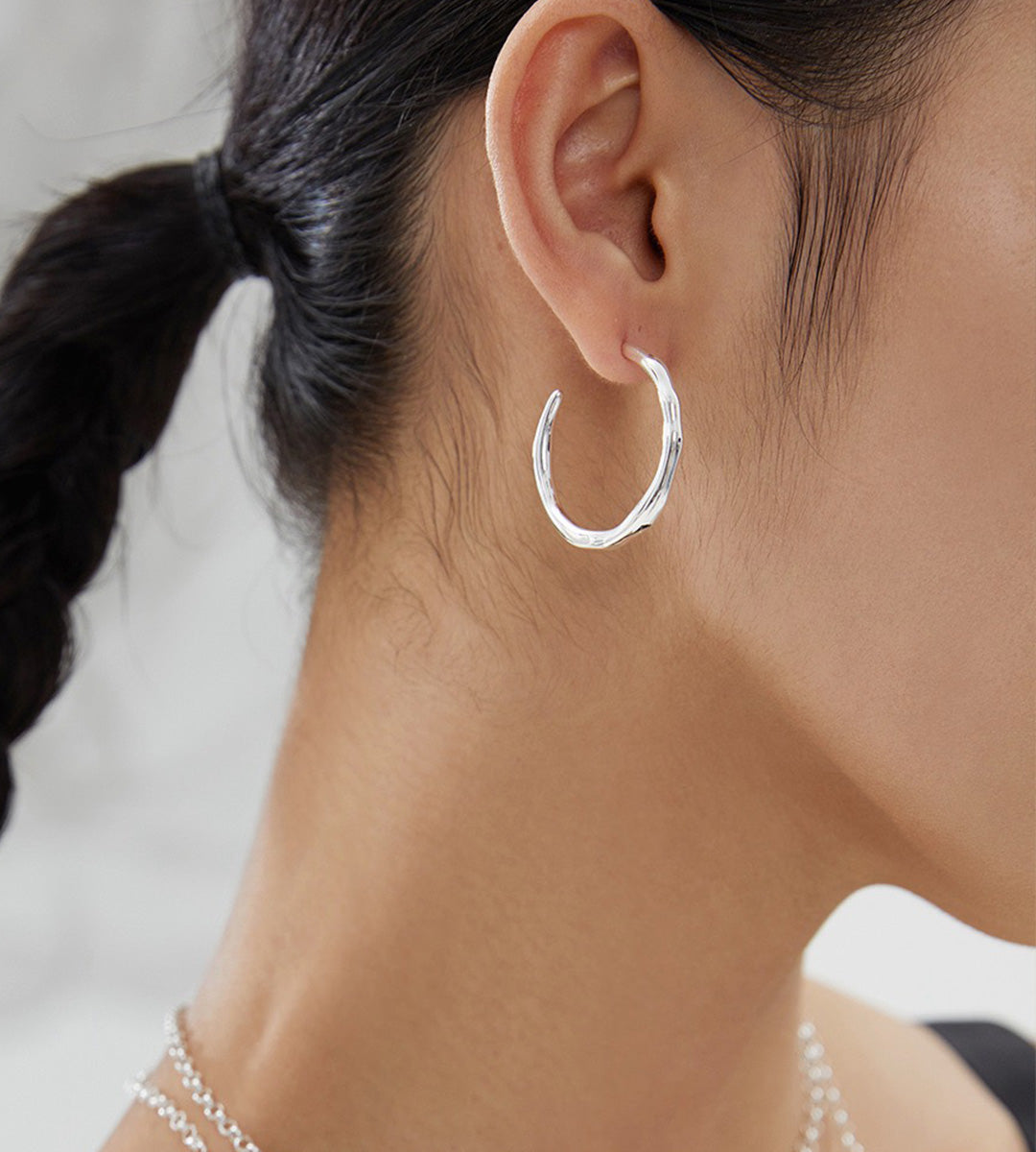 A close-up of a woman's ear wearing a textured 18K white-gold-plated S925 silver huggie hoop earring, with her dark hair tied back in a ponytail.