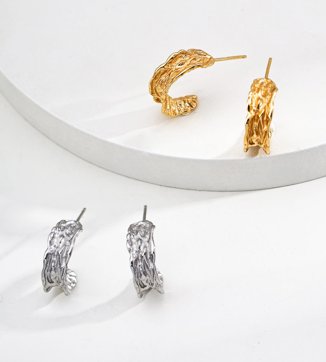 Two pairs of textured hoop-style earrings are displayed on a clean white surface, including two 18K gold-plated hoops on a raised white platform and one pair of 18K white-gold hoops below.
