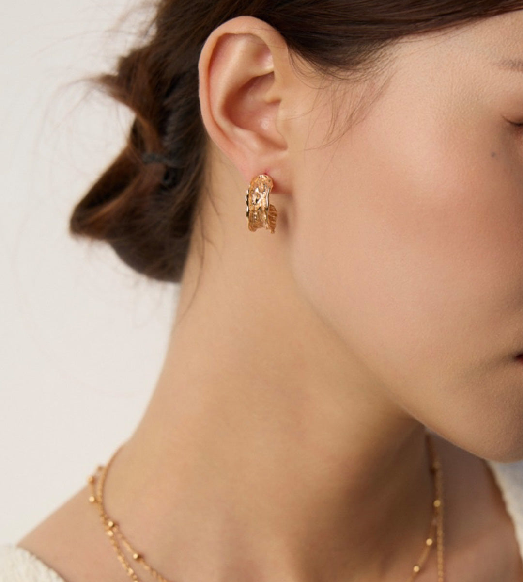A close-up lifestyle shot of a model wearing a delicate 18K gold-plated S925 silver organic textured hoop earring, along with a fine gold chain necklace against a clean, light background.