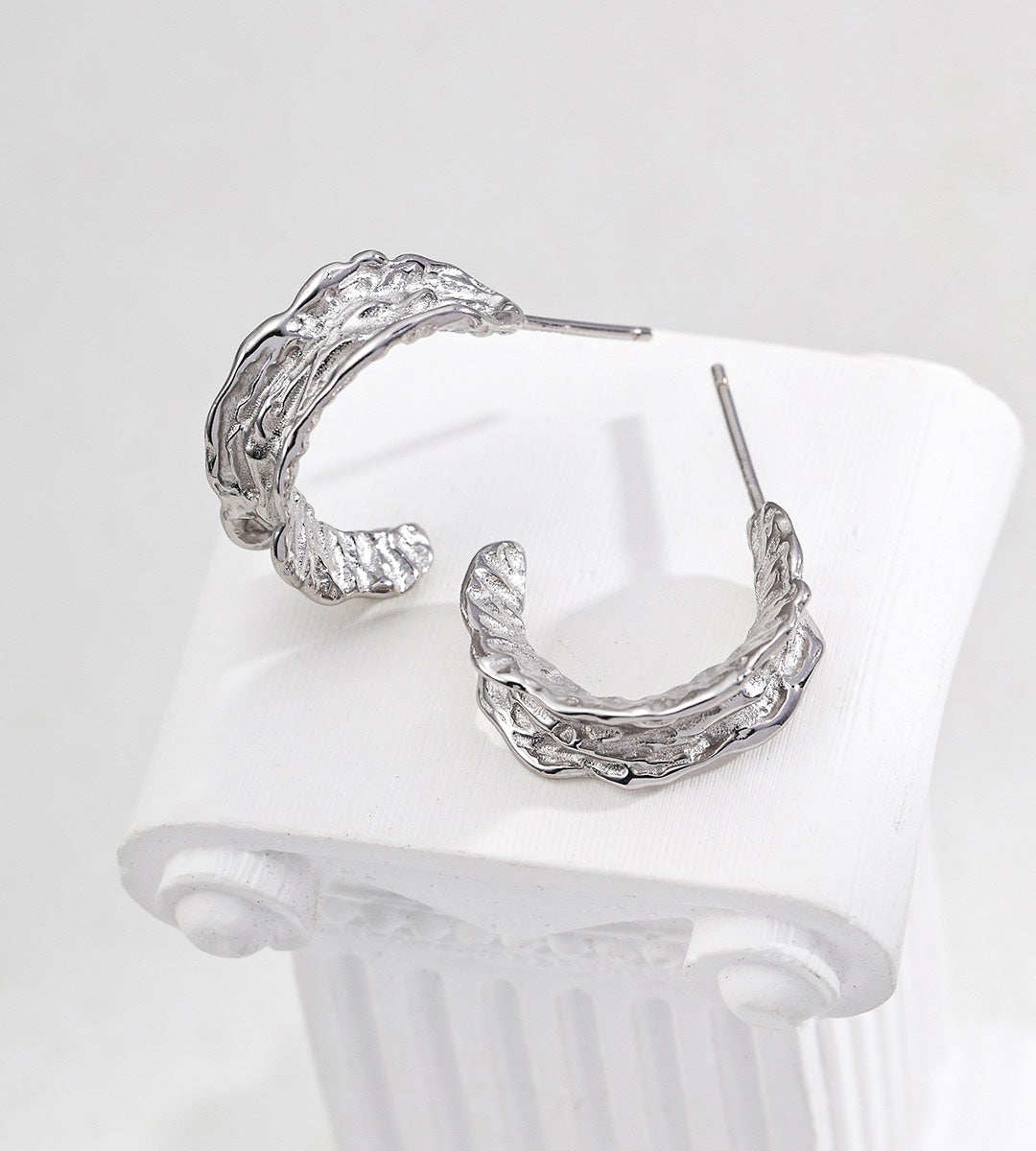 A pair of textured 18K white-gold-plated S925 silver hoop earrings displayed on a white ribbed surface against a light gray background.