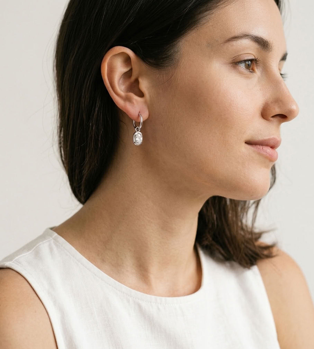 A close-up side profile of a woman wearing a delicate silver huggie hoop earring with a small clear teardrop gemstone dangle, against a plain light background.