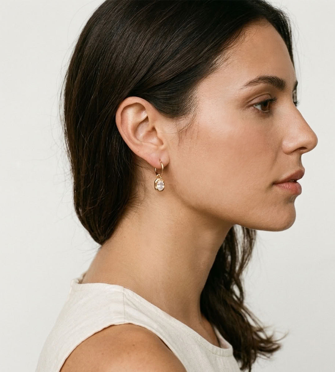 A woman in profile wearing a delicate gold huggie hoop earring with a small clear teardrop gemstone, shown against a plain light background.