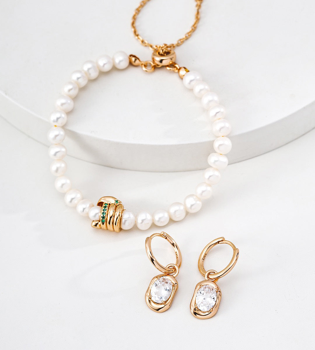An 18K gold-plated S925 silver and pearl jewelry set featuring a pearl bracelet with a gold clasp and decorative bead, matching 18K gold-plated S925 silver hoop earrings with zirconia stones, arranged on a white curved pedestal.