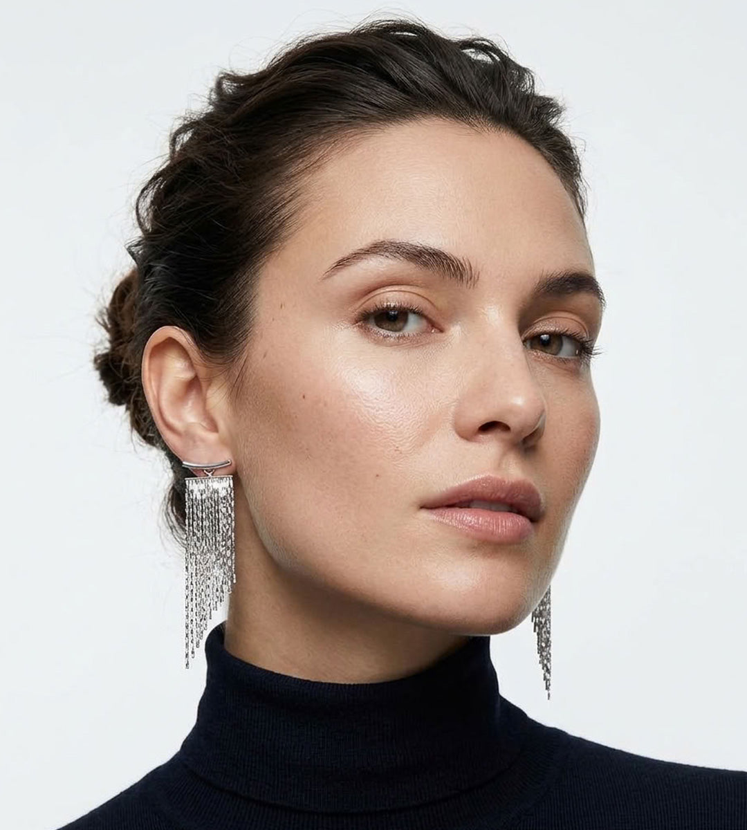 A close-up portrait of a woman wearing long, dangling 18K white-gold-plated earrings with a textured, chain-like fringe design, paired with a dark navy turtleneck sweater against a light gray background.
