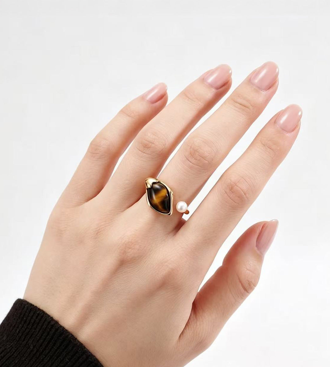 A hand wearing a gold ring with a marquise-cut brown tiger's eye stone and a small white pearl.