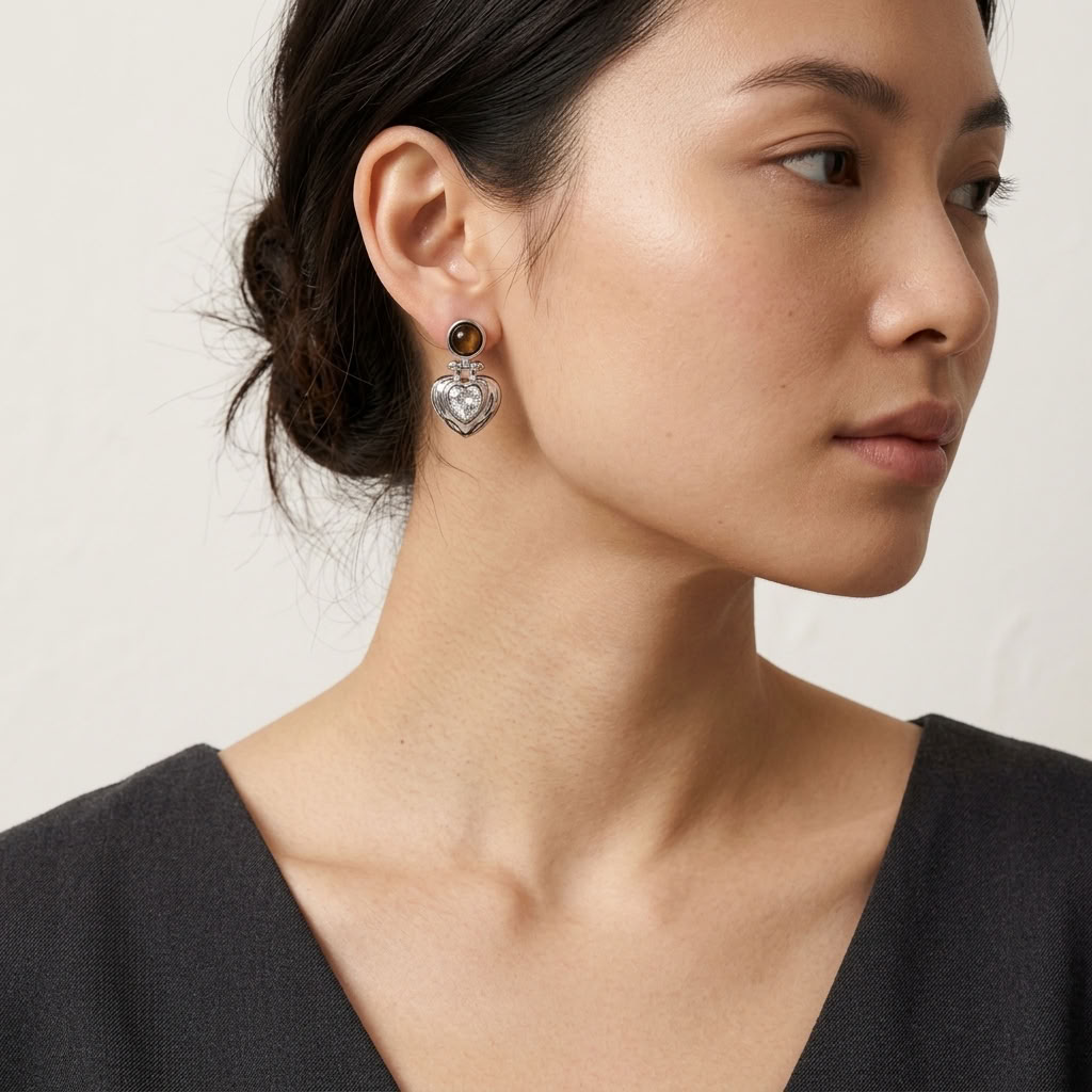 A close-up side profile of a woman wearing a silver heart-shaped dangle earring with a tiger stone and clear crystal detailing, paired with a black V-neck top against a light beige background.