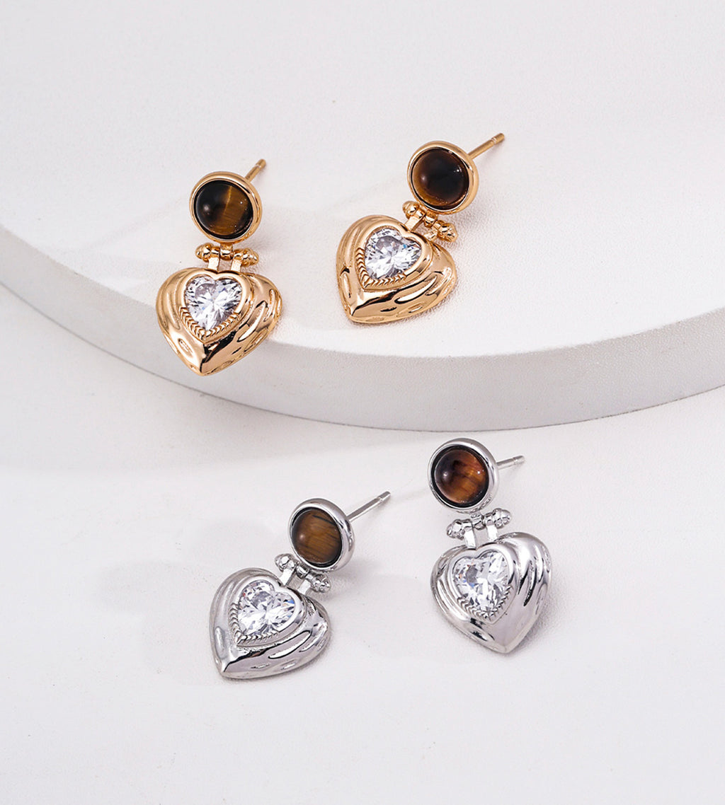 Three pairs of dangling heart stud earrings are arranged on a white curved platform and white surface. Each earring has a small round brown stone above a larger heart-shaped setting with a clear central stone.