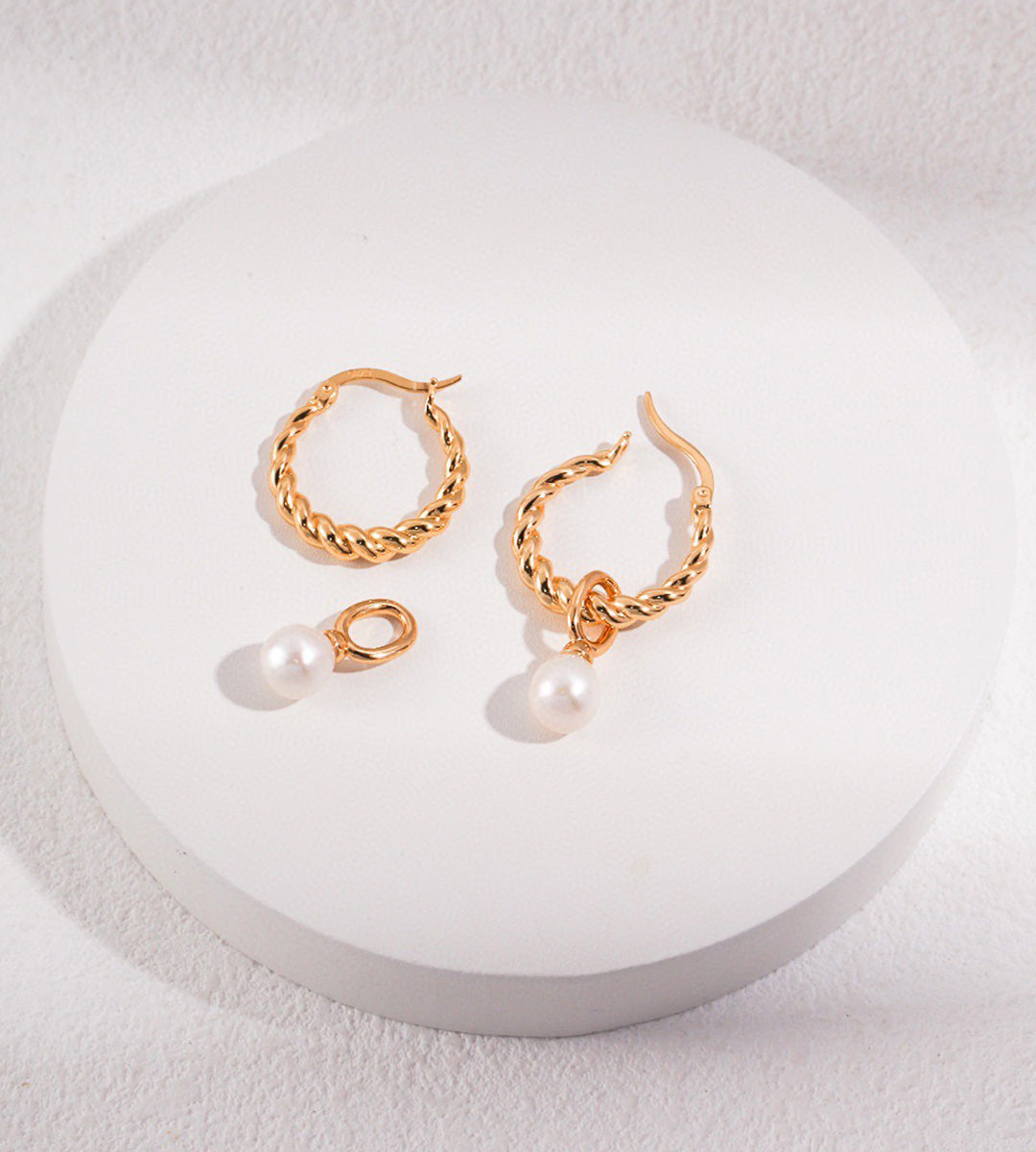 A pair of 18K gold-plated twisted hoop earrings with dangling pearl charms, displayed on a round white pedestal against a textured white background.