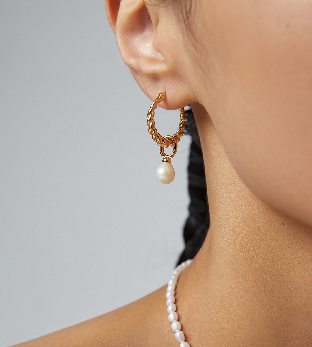 A close-up of a model's ear wearing an 18K gold-plated hoop earring with a dangling white pearl, alongside a matching pearl necklace against a neutral gray background.