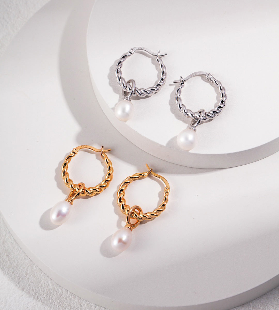 Two pairs of twisted hoop earrings with dangling pearl drops, shown on a white curved display surface. One pair is 18K white-gold-plated, and the other is 18K gold-plated