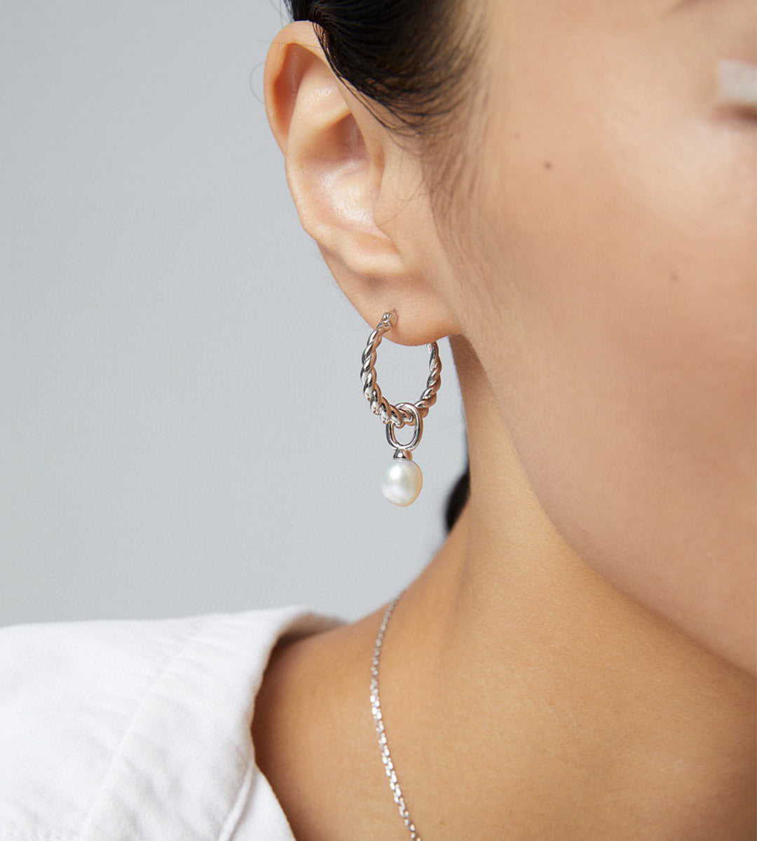 A close-up of a model's ear wearing an 18K white-gold-plated hoop earring with a dangling white pearl, paired with a delicate silver chain necklace.