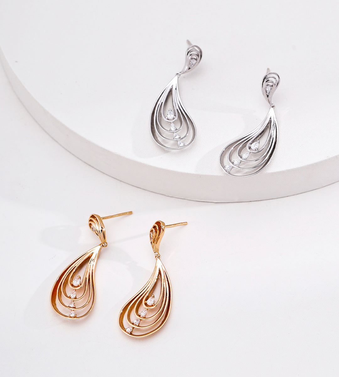 Three teardrop-shaped dangle earrings with a layered, swirling openwork design are displayed on a white surface and a white curved riser. Two earrings are 18K white-gold-toned, and one is 18K gold-plated, each featuring small cubic zirconia stone accents