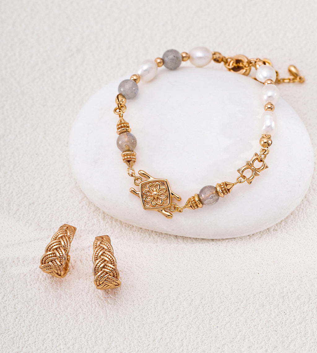 18K gold-plated S925 silver beaded bracelet with gray stone beads, white pearl beads, and a central decorative charm, displayed on a white stone with matching gold-plated earrings on a textured cream background.