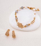 18K gold-plated S925 silver beaded bracelet with gray stone beads, white pearl beads, and a central decorative charm, displayed on a white stone with matching gold-plated earrings on a textured cream background.