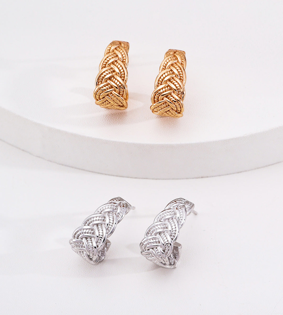 Two pairs of braided hoop earrings displayed on a white surface, one pair in 18K gold-plated S925 silver metal and one pair in 18K white-gold-plated S925 silver, with the gold pair positioned higher on a white curved platform.