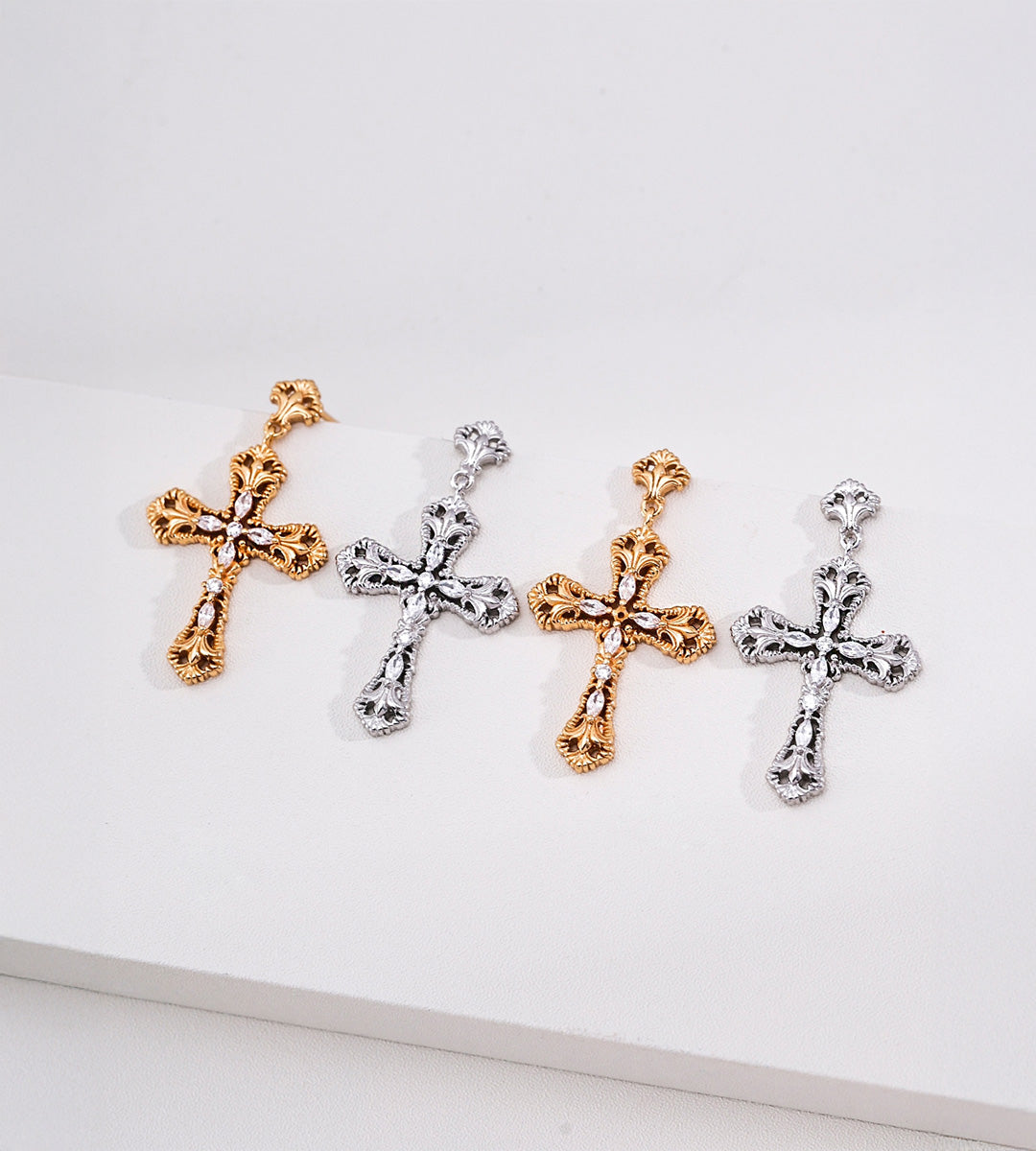 Four ornate cross drop earrings in alternating gold and silver tones, with scrollwork and clear stone accents.