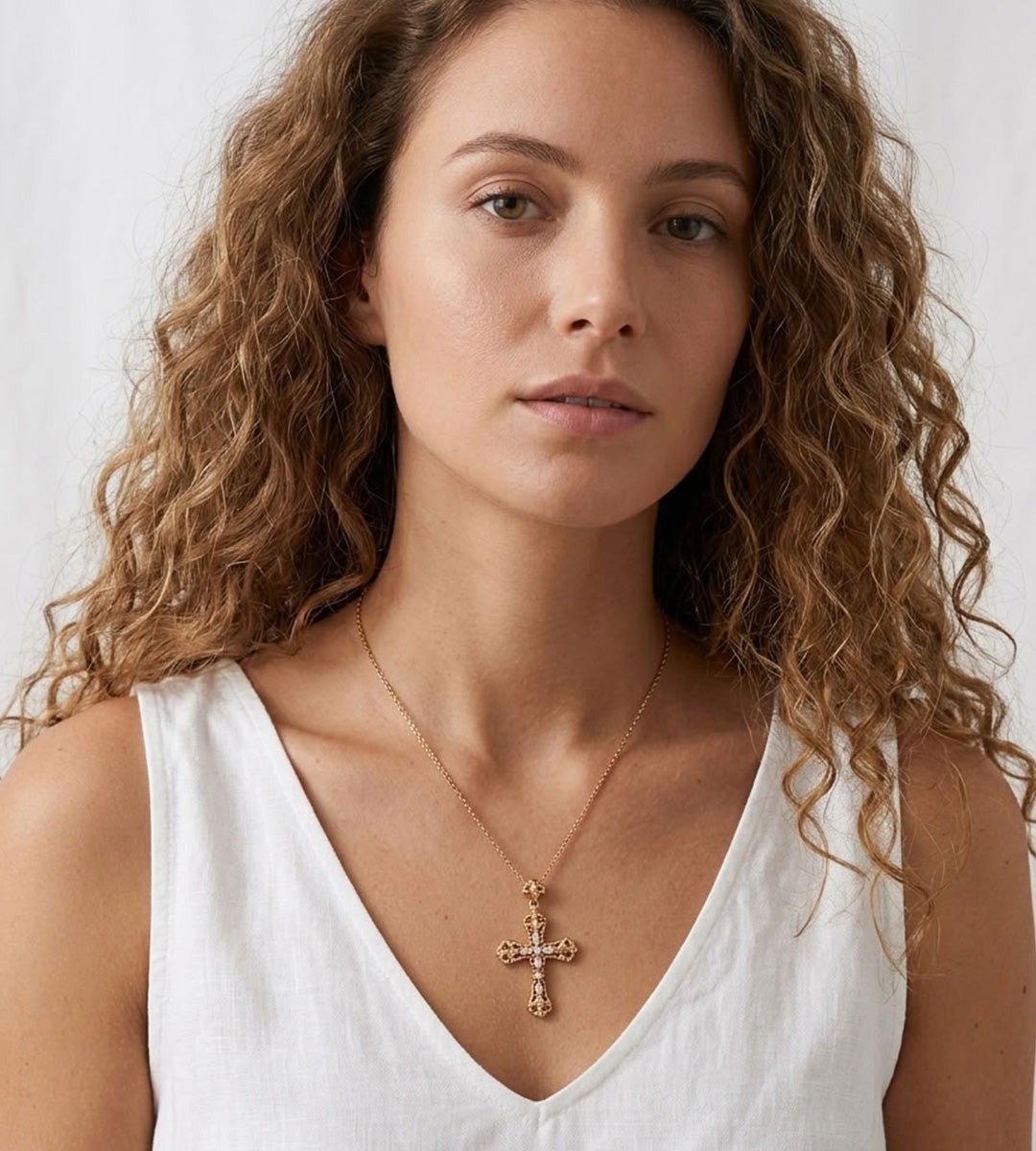 A close-up of a woman with curly brown hair wearing a delicate gold cross pendant necklace over a white sleeveless top, set against a soft white background.