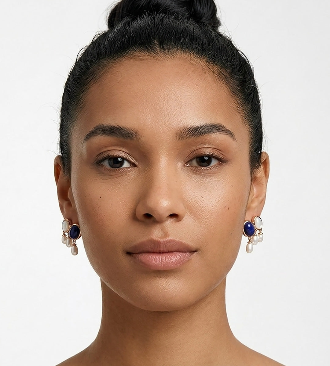 A close-up portrait of a woman with her hair in a bun, wearing a pair of 18K gold-plated S925 Silver drop earrings with a lapis lazuli oval stone, mother-of-pearl, and three small natural freshwater AAAA-grade pearls. BUCKLE Collective