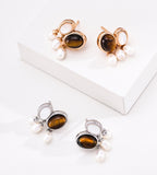 Four pairs of drop earrings displayed on a white surface, including two gold and two silver-toned pairs, each featuring a tiger's eye stone and small white pearl drops.